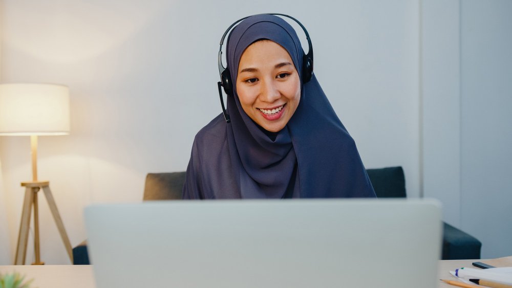 Smiling online Islamic tutor wearing a headset and hijab, teaching a personalized one-to-one Arabic and Quran lesson on a computer.